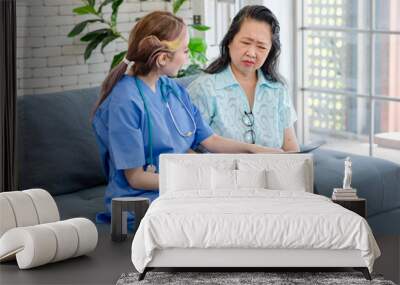 Asian young professional female doctor in blue hospital uniform with stethoscope sitting on sofa couch holding spine xray film discussing explaining to old chubby retirement patient in living room Wall mural