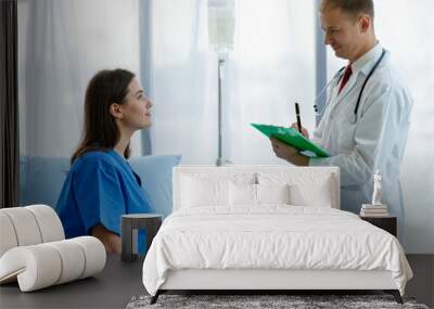 At hospital woman caucasian patient sitting on bed and caucasian doctor standing near her, both are smiling happily. Doctor man carrying stethoscope on neck and hold a note Wall mural