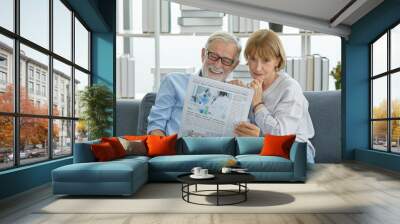 Caucasian old senior elderly grandparent grey bearded hair husband and pretty wife sitting cuddling together on cozy sofa in living room at home in morning reading funny news on daily newspaper Wall mural