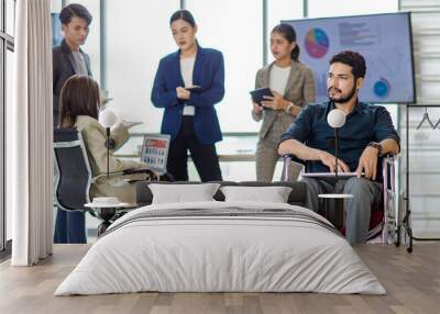 Indian bearded disabled handicapped businessman sitting working with laptop computer on wheelchair while Asian male and female colleagues hold tablet talking discussing together in blurred background Wall mural