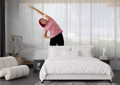 Lively Asian woman on pink shirt, black trousers, red scarf as breast cancer patient, standing on carpet and exercise by side body bending and stretch hand up to strengthen health as disease cure Wall mural