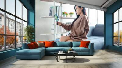 Millennial Asian beautiful pretty female model in casual outfit big brown warm sweater sitting on cozy sofa couch holding handheld mirror looking at her reflection adjusting hairstyle in living room Wall mural