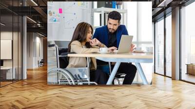 Millennial Indian male businessman manager mentor in formal suit talking helping disabled handicapped female employee sit on wheelchair working with laptop computer on table in office meeting room Wall mural