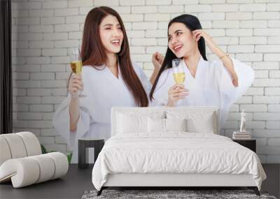 Millennial two Asian beautiful drunk girlfriends in white clean bathrobes celebrating party relaxing resting holding drinking tall glasses of golden sparkling champagne in spa massage studio resort Wall mural