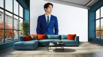 Portrait closeup shot of Asian young handsome happy intelligence college teen student wears blue formal suit and necktie standing hold hands in pants pockets look outside in front of white wall Wall mural