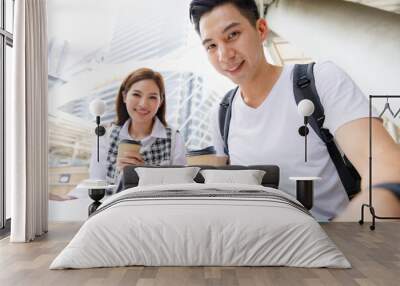 Portrait of cute smiling young Asian couple tourists sitting on stair while holding paper city map and coffee disposable cup together with a blurred city building background. Selective focus at woman Wall mural