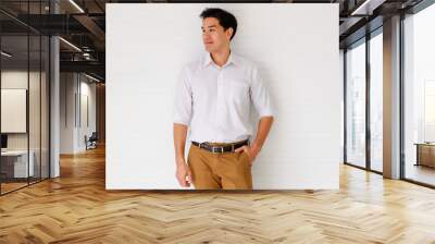 Portrait shot of young adult Asian confident man with cute smiling wearing a formal white long sleeve shirt and brown pants with hand in pocket posturing isolated with white background in the studio Wall mural