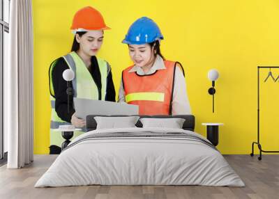 Studio shot of two Asian female professional engineering manager foreman wearing hard helmet and reflective safety vest holding paperwork document file folder discussing together on yellow background Wall mural