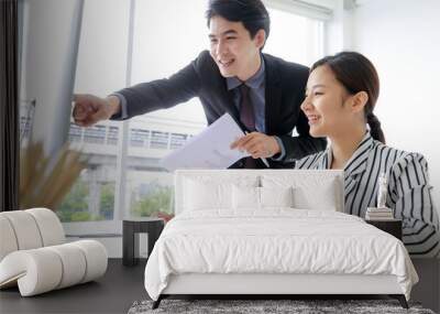 Two asian business man and woman smiling with happiness, discussing about their project and brainstorming creative idea together in the indoor office or workplace while looking, pointing at computer. Wall mural