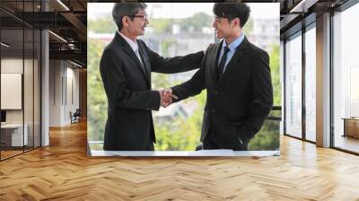 Two Asian businessmen wearing a formal suit, looking at each other eyes with pleasure smiling. The senior manager congratulates a successful subordinate by touching his shoulders and shake the hand Wall mural