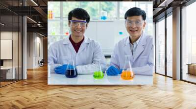 Two Asian male scientists in white lab coat safety goggles rubber gloves working discussing talking together about colorful liquid samples in glass flasks in scientific experiment on laboratory table Wall mural