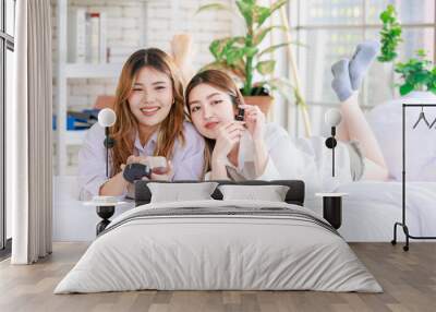 Two happy beautiful Asian female smile lying relaxed on a white bedroom, putting makeup. Attractive woman having fun together trying beauty products, Concept teenage or girlfriends lifestyle at home. Wall mural