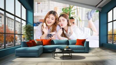 Two happy beautiful Asian female smile lying relaxed on a white bedroom, putting makeup. Attractive woman having fun together trying beauty products, Concept teenage or girlfriends lifestyle at home. Wall mural