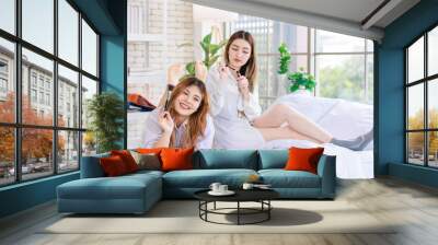 Two happy beautiful Asian female smile lying relaxed on a white bedroom, putting makeup. Attractive woman having fun together trying beauty products, Concept teenage or girlfriends lifestyle at home. Wall mural