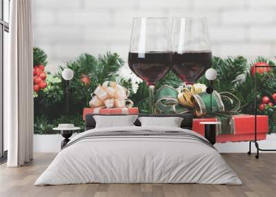 Two red wine glasses set next to red gift boxex on snowy table prepare for celabraion, decorate with pine branches and holly berries. Blurred white brick wll copy space at top part Wall mural