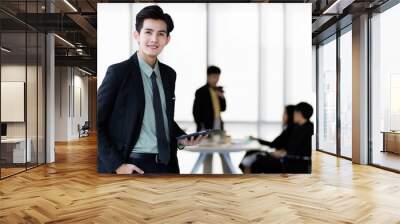 Young and handsome Asian businessman holding tablet computer  standing pose in modern office with self-confidence and success gesture with other colleagues blur in background Wall mural