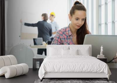 Young cute and beautiful secretary in company sitting beside white hard safety hat and using labtop notebook computer to work with two businessmen talking in blur background Wall mural