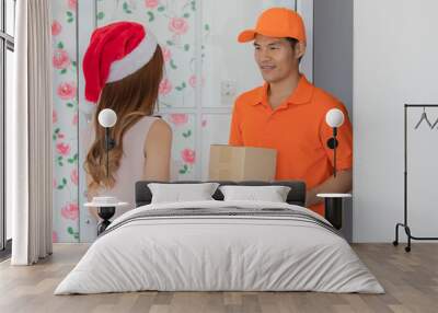 Young female wearing red and white santa hat and pink sleeveless dress stand receiving boxes of package from delivery service asian man wearing orange shirt and cap at glass door in front of her home. Wall mural