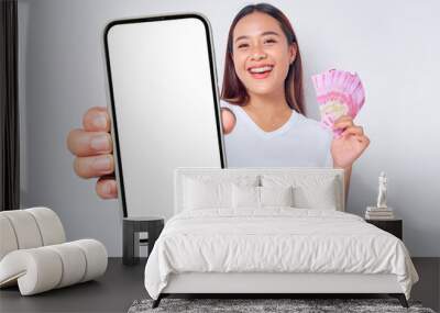 Cheerful young blonde woman girl Asian wearing casual white t-shirt  showing mobile phone with blank screen and holding money rupiah banknotes isolated on white background. people lifestyle concept Wall mural