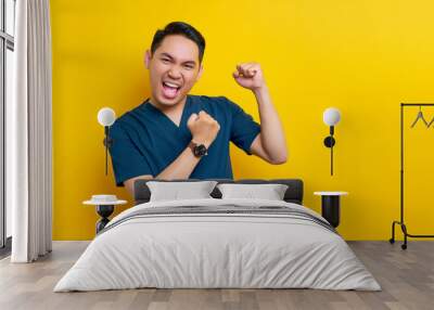 Excited professional young Asian male doctor or nurse wearing a blue uniform standing confidently and celebrating success isolated on yellow background. Healthcare medicine concept Wall mural