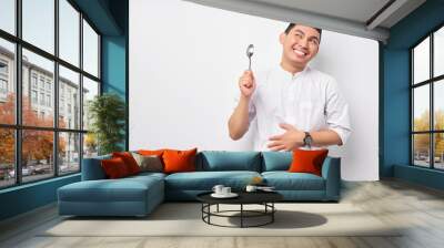 Feeling hungry young Asian Muslim man touching hand on the stomach while holding spoon, wait for delicious food to break fast isolated on white background. People religious Islamic lifestyle concept Wall mural