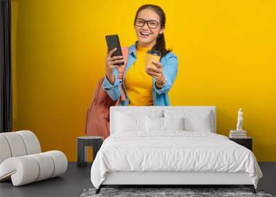 Portrait of cheerful young Asian woman student in denim clothes with backpack holding cup of coffee and smartphone isolated on yellow background Wall mural