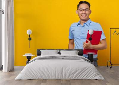 Portrait of handsome smiling young man in glasses holding document folder isolated on yellow background Wall mural