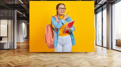 Portrait of smiling young Asian woman student in casual clothes with backpack holding book and pointing aside with finger isolated on yellow background. Education in college university concept Wall mural