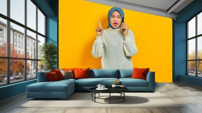 Shocked young asian woman in white sweater and hijab holding mobile phone and pointing fingers up isolated over yellow background. People islam religious concept Wall mural