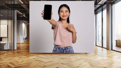 Smiling happy fun beautiful young Asian woman in a casual t-shirt pointing index finger at mobile phone with blank screen isolated on white background Wall mural