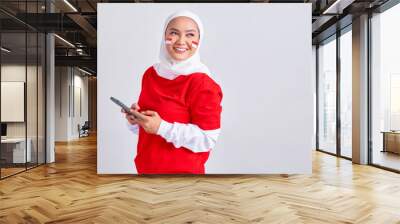 Smiling young Asian muslim woman in red white t-shirt using mobile phone and looking aside at copy space isolated on white background. Indonesian independence day on 17 august concept Wall mural