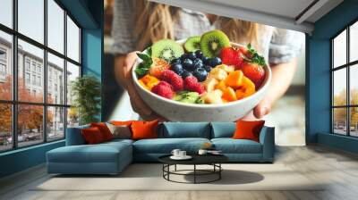 A vibrant bowl of mixed fruit held by a person, showcasing fresh strawberries, blueberries, kiwi, and oranges in a cozy kitchen setting. Wall mural