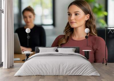 Two women are engaged in a professional setting, one focused in front, while the other works on a laptop. Wall mural