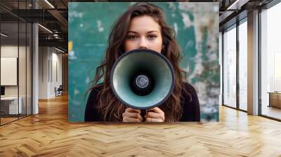 A young woman with long brown hair holds a megaphone to her mouth in front of a weathered green wall. Wall mural