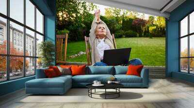 adult woman stretches arms overhead sitting on terrace with laptop working remotely in afternoon from home Wall mural