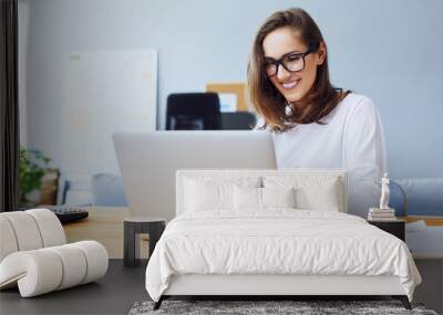 Attractive cheerful young businesswoman working on laptop and smiling while sitting at her desk in bright modern office Wall mural