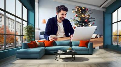 Cheerful man working from home with laptop and phone during Christmas Wall mural