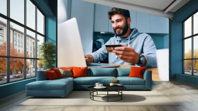 Cheerful young man paying bills online with credit card and laptop Wall mural