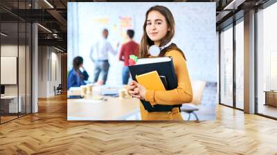 Female student standing in classroom holding books. Portrait of young woman looking at camera indoors Wall mural