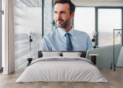 Handsome businessman standing with laptop, looking throught the window in modern office Wall mural