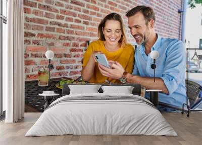 Happy couple sharing smartphone while out in the city eating lunch at outdoors cafe Wall mural