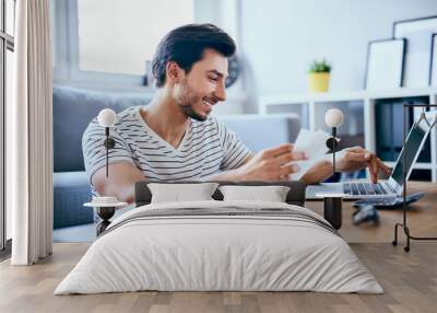 Happy man paying bills on his laptop in living room Wall mural