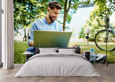 Happy young businessman using laptop and phone while sitting in the park during lunch break Wall mural