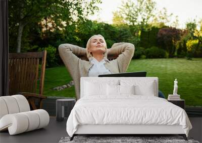 Mature woman with eyes closed relaxing on backyard garden terrace sitting on a chair with laptop Wall mural