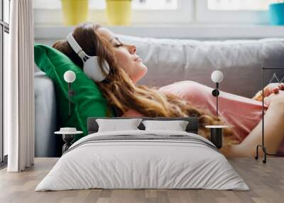 Portrait of a beautiful young woman lying on sofa with headphones on and closed eyes, relaxing Wall mural