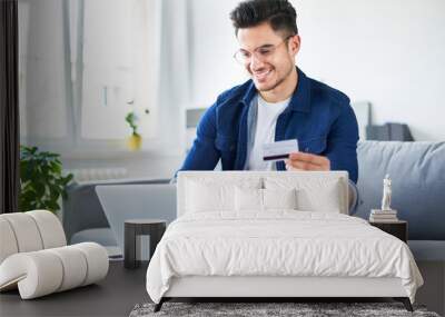 portrait of handsome young man paying online with credit card and laptop while sitting at home Wall mural