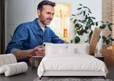 Smiling adult man using laptop and phone while freelancing at home Wall mural