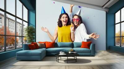 two cheerful party girls raising arms and laughing in birthday hats Wall mural