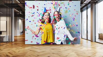 Two ecstatic young women laughing and raising arms in birthday hats with confetti fyilng around them Wall mural