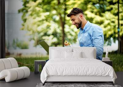 Young cheerful businessman sitting in the park with sandwich and laptop Wall mural